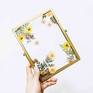 May include: A gold-toned picture frame with a clear glass front and a stand on the back. Dried flowers and leaves are pressed inside the frame.