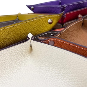 Op de afbeelding: Een set lederen trays in verschillende kleuren, waaronder wit, geel, bruin, oranje, rood en paars. De trays zijn gemaakt van een getextureerd leer en hebben een gestikte rand.