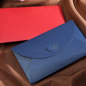 May include: Two envelopes, one red and one navy blue, rest on a brown satin surface. The navy envelope has a heart-shaped cutout on the flap. The envelopes are likely for invitations or special occasions.