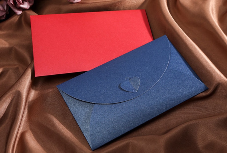 May include: Two envelopes, one red and one navy blue, rest on a brown satin surface. The navy envelope has a heart-shaped cutout on the flap. The envelopes are likely for invitations or special occasions.
