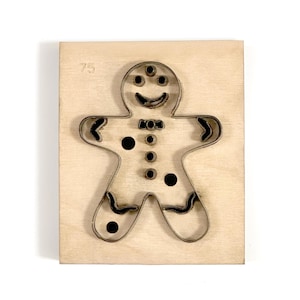 May include: A gingerbread man cookie cutter made of metal, set on a light-colored wooden base. The cutter has a smiling face, bow tie, and button details. The number "75" is printed on the wood.