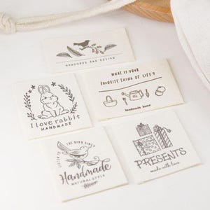 Puede incluir: Cinco etiquetas rectangulares de tela con ilustraciones y texto en tinta negra. Los diseños incluyen un pájaro, un conejo, cajas de regalo y las frases "Handmade and Design", "I love rabbit Handmade", "What is your favorite thing of life? handmade home", "Presents made with love", y "Handmade Natural Style".