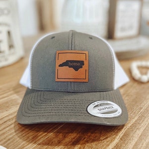 Może przedstawiać: Szara i biała czapka trucker z brązową skórzaną naszywką przedstawiającą zarys Karoliny Północnej i napis "home". Czapka ma biały siateczkowy tył i szary daszek.