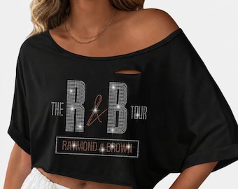 Op de afbeelding: Een zwarte crop top met korte mouwen, een wijde halslijn en een kleine horizontale opening bij de kraag. De voorzijde is voorzien van een strass-afbeelding met de tekst THE R & B TOUR en RAYMOND & BROWN in een rechthoekig kader.