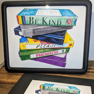 May include: A stack of vintage VHS tapes with colorful labels. The top tape has the text "Be Kind" and the bottom tape has the text "Please Rewind".