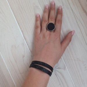 May include: A black leather bracelet with two straps, worn on a person's wrist. The bracelet is made of two thin strips of leather, each with a textured surface. The person is also wearing a large, round ring with a black centre and a silver band.