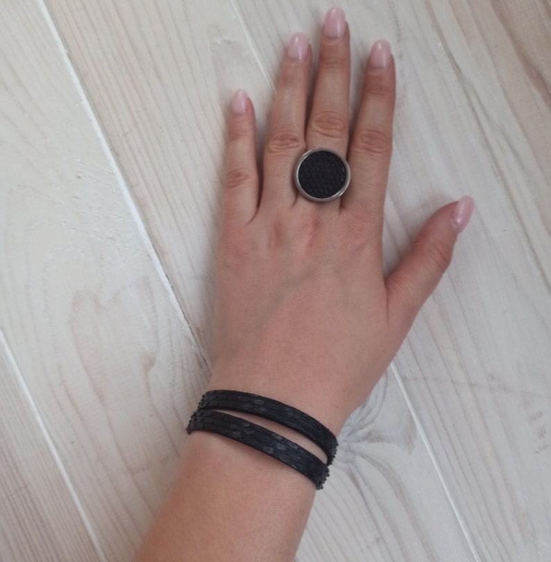 May include: A black leather bracelet with two straps, worn on a person's wrist. The bracelet is made of two thin strips of leather, each with a textured surface. The person is also wearing a large, round ring with a black centre and a silver band.