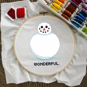 May include: A white fabric with a wooden embroidery hoop. The hoop contains a cross-stitch design of a snowman with the text "WONDERFUL."  A spool of red embroidery floss is on the fabric.