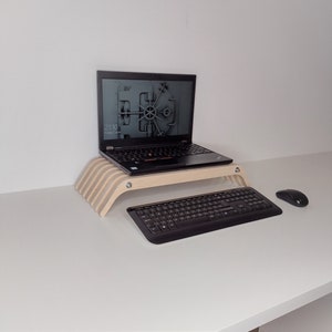 May include: A laptop computer with a black keyboard and a wooden laptop stand. The laptop is on a white desk with a black wireless keyboard and a black mouse.