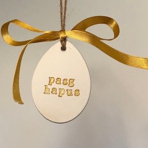 May include: White ceramic egg-shaped ornament with a gold ribbon and the text "pasg hapus" in gold lettering.