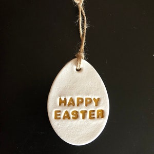 Könnte beinhalten: Weißer Keramik-Eieranhänger mit goldener Aufschrift "Frohe Ostern" und einem Bindfaden zum Aufhängen.