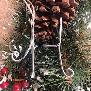 Könnte beinhalten: Silberner Draht-Anhänger in Form des Buchstabens H, der an einer braunen Schnur an einem grünen und weißen künstlichen Weihnachtsbaum hängt.