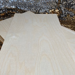 Op de afbeelding: Twee onafgewerkte, lichtgekleurde houten planken met een stervormige bovenkant. De planken zijn op elkaar gestapeld, waarbij de bovenste plank iets schuin staat. De achtergrond is een sprankelende, met pailletten bezette stof.