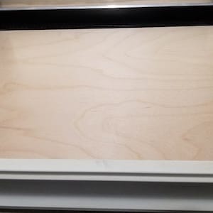 Op de afbeelding: Een lichtgekleurd, rechthoekig houten paneel, waarschijnlijk multiplex, is geplaatst in een zwart frame. De houtnerf is zichtbaar en het paneel is omgeven door een witte rand. De afbeelding is een close-up.