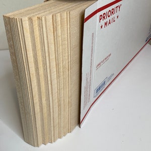 Op de afbeelding: Een stapel onafgewerkte, lichtgekleurde houten planken, gedeeltelijk in een witte en rode Priority Mail flat rate doos gestoken. De planken zijn netjes opgestapeld en tonen hun randen. De doos is open en onthult de inhoud.