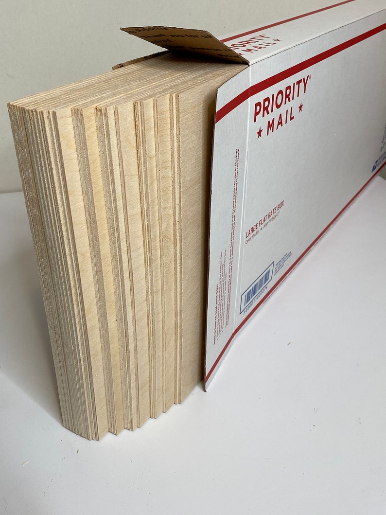 Op de afbeelding: Een stapel onafgewerkte, lichtgekleurde houten planken, gedeeltelijk in een witte en rode Priority Mail flat rate doos gestoken. De planken zijn netjes opgestapeld en tonen hun randen. De doos is open en onthult de inhoud.