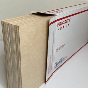 Op de afbeelding: Een stapel lichtgekleurde houten planken, gedeeltelijk in een witte en rode Priority Mail flat rate box gestoken. De planken zijn netjes opgestapeld en tonen hun randen. Op de doos staat "PRIORITY MAIL".
