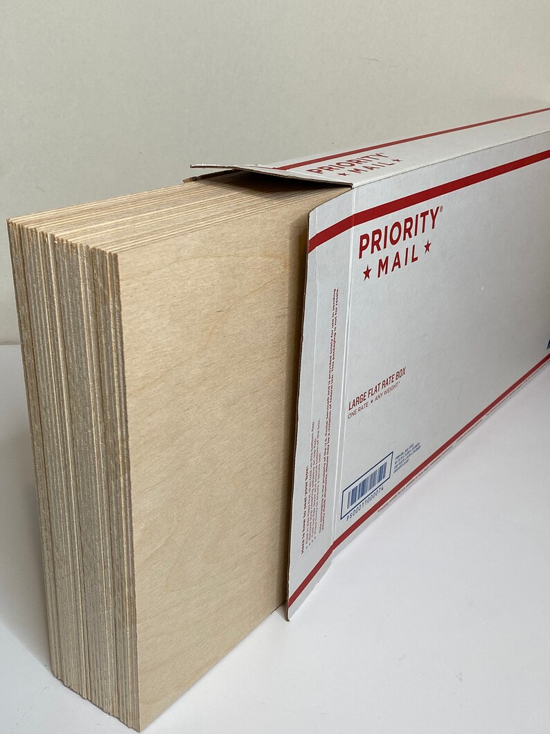 Op de afbeelding: Een stapel lichtgekleurde houten planken, gedeeltelijk in een witte en rode Priority Mail flat rate box gestoken. De planken zijn netjes opgestapeld en tonen hun randen. Op de doos staat "PRIORITY MAIL".