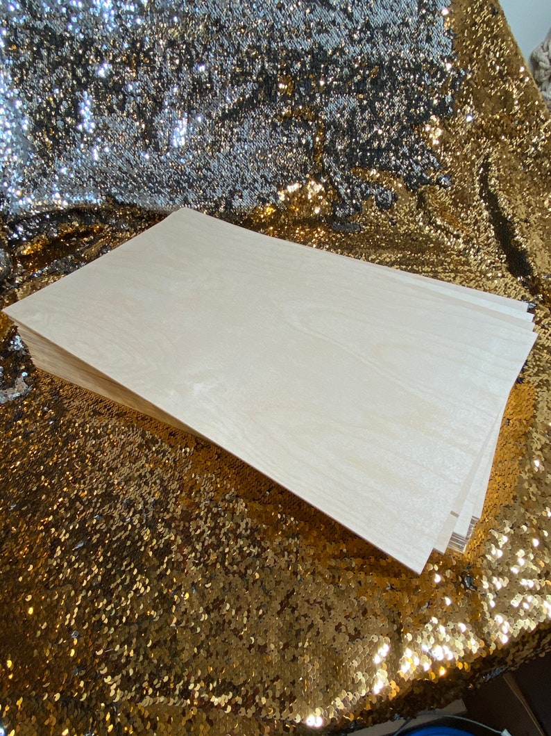 Op de afbeelding: Een stapel lichtgekleurde houten planken, mogelijk multiplex of een vergelijkbaar materiaal, met zichtbare houtnerf. De planken zijn netjes opgestapeld en tonen hun randen. De achtergrond is een gouden paillettenstof.
