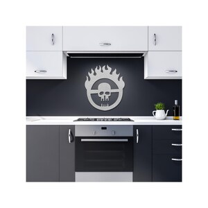 May include: A silver metal wall art piece featuring a skull within a flaming circle design. The artwork is mounted on a dark gray wall in a kitchen setting, above a stove and oven. The kitchen has white and dark gray cabinets.