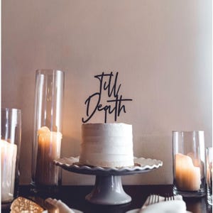 May include: A white cake with a black acrylic cake topper that says "Till Depth". The cake is on a white cake stand with a scalloped edge. There are three clear glass cylinders with candles in them on the table.
