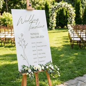Op de afbeelding: Een wit trouw-tijdschema bord op een houten ezel met olijfgroene tekst en een florale lijntekening. Het bord toont de datum maandag 24 juni 2024 en een schema van evenementen van 06.00 tot 11.00 uur, versierd met witte bloemen en groen.