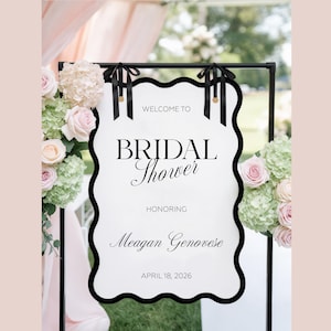 May include: A white scalloped sign with a black border hanging from a black metal frame by two black ribbons. The text reads: WELCOME TO BRIDAL Shower HONORING Meagan Genovese APRIL 18, 2026. The sign is decorated with pink roses and green hydrangeas.