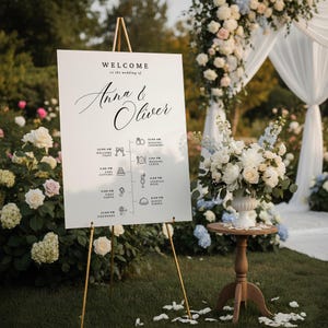 May include: A wedding sign on a gold easel, welcoming guests to the wedding of Anna & Oliver. The sign includes a schedule with icons. A floral arrangement and archway with white and blue flowers are visible in the background.