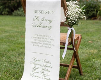 May include: A wooden folding chair draped with a white fabric sign featuring the text RESERVED In loving Memory FOR THOSE WHO CANNOT BE HERE TODAY BUT ARE FOREVER PRESENT IN OUR HEARTS, TODAY AND ALWAYS, followed by five names and a floral bouquet.