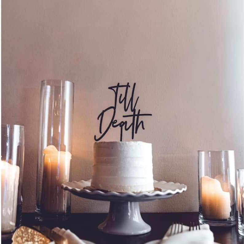 May include: A white cake with a black acrylic cake topper that says "Till Depth". The cake is on a white cake stand with a scalloped edge. There are three clear glass cylinders with candles in them on the table.