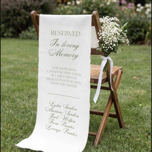 May include: A wooden folding chair draped with a white fabric memorial sign featuring the text RESERVED In loving Memory FOR THOSE WHO CANNOT BE HERE TODAY BUT ARE FOREVER PRESENT IN OUR HEARTS, TODAY AND ALWAYS and five names, with baby's breath.