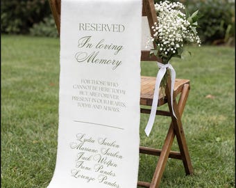 May include: A wooden folding chair draped with a white fabric memorial sign featuring the text RESERVED In loving Memory FOR THOSE WHO CANNOT BE HERE TODAY BUT ARE FOREVER PRESENT IN OUR HEARTS, TODAY AND ALWAYS and five names, with baby's breath.