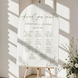 Puede incluir: Un plano de asientos de boda blanco con una parte superior arqueada, sostenido por un caballete de madera. El gráfico presenta las palabras "find your seat" y los nombres "Jonathan + Juliana" con la fecha "June 20, 2026". Los nombres se enumeran debajo de los números de mesa.