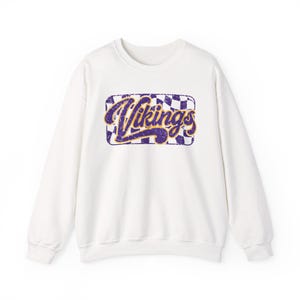 May include: A white long-sleeve crewneck sweatshirt featuring a central graphic with a purple and white checkered background. The word Vikings is printed in a distressed purple script font with a yellow outline, centered within the design.