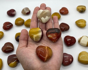 May include: A collection of heart-shaped polished stones in shades of red, yellow, and brown. The stones are arranged on a white surface, with one hand holding a few of the stones. A gold bracelet is worn on the wrist.