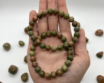 May include: Two green and red jasper stone bracelets with round beads. The bracelets are stacked on a person's wrist.