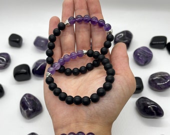 May include: Three stacked bracelets with black matte beads and amethyst gemstone beads. The bracelets have silver accents.