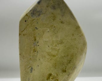 May include: A polished stone sculpture with a pointed top and a curved side. The surface is a pale, creamy yellow color with natural mottled patterns, dark speckles, and subtle veining throughout the dense, smooth, and solid mineral structure.