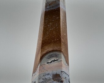 Könnte beinhalten: Ein hoher, vielseitiger Steinobelisk mit spitzer Spitze. Der Obelisk weist Bänder in Braun-, Grau- und Rosatönen mit einem strukturierten, geschichteten Aussehen auf. Die Basis hat eine sechseckige Form.
