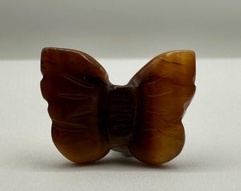 May include: A carved brown gemstone butterfly figurine with a smooth, polished surface and subtle natural banding. The wings feature delicate etched lines, and the central body is defined by vertical grooves, showcasing a rich, warm amber tone.