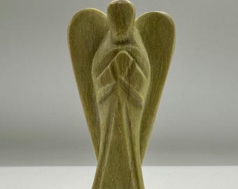 May include: A light olive green stone angel figurine with a smooth, polished finish. The figure features stylized, elongated wings and hands held together in a prayer position at the center of the chest, with a simple, flowing robe design.