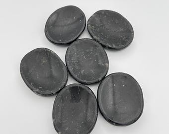 May include: Six black oval-shaped polished stones, also known as palm stones, are arranged in a circle on a white surface.