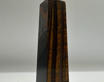 May include: A polished tiger's eye crystal tower with a pointed top. The stone features vertical bands of dark brown and golden-brown chatoyant fibers, creating a striped pattern that runs along the length of the four-sided obelisk shape.