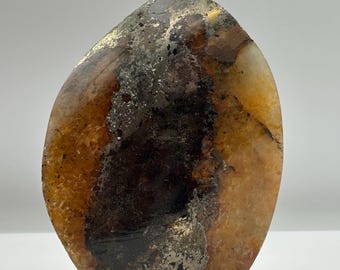 May include: A polished, oval-shaped mineral specimen featuring a central band of dark, metallic brown and black inclusions flanked by translucent, amber-colored crystalline sections with visible internal fractures and golden pyrite flecks.