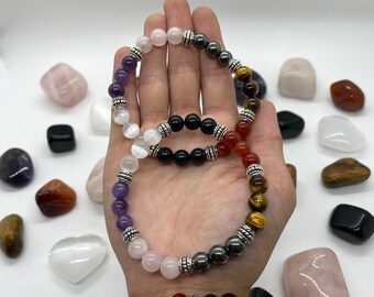 May include: A set of seven gemstone bracelets with silver beads. The bracelets are stacked on a person's wrist and feature amethyst, rose quartz, tiger's eye, carnelian, and hematite beads.