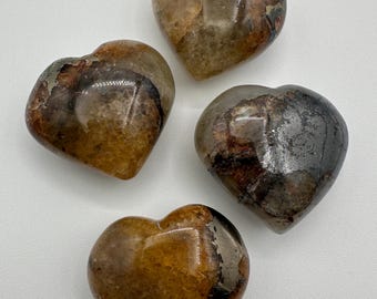 May include: Four polished heart-shaped stones featuring a mix of amber, brown, and dark gray tones. Each stone has a smooth, glossy surface with visible natural inclusions, varying patterns of dark mineral deposits, and translucent sections.