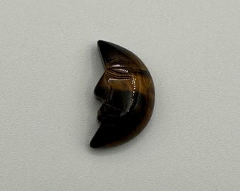 May include: A crescent moon carving made from polished tiger's eye gemstone. The stone features natural bands of golden-brown and dark brown colors. The carving depicts a human-like face in profile with closed eyes, a nose, and a mouth.