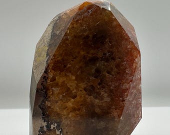 May include: A polished, upright crystal specimen featuring a deep amber and reddish-brown color palette. The stone has a faceted, geometric shape with a textured, crystalline interior surface and smooth, dark edges that catch the light.