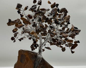 May include: A decorative tree sculpture featuring a thick, brown, natural stone base. The trunk and branches are crafted from twisted silver-colored wire, with numerous small, polished, brown and gold tiger's eye gemstone chips attached.