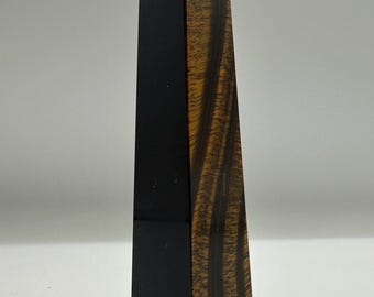 May include: A tall, four-sided obelisk sculpture featuring a smooth, solid black side and a contrasting side with a natural, wood-like grain pattern in shades of dark brown and golden tan. The object has a pointed, pyramid-shaped top and a flat base.